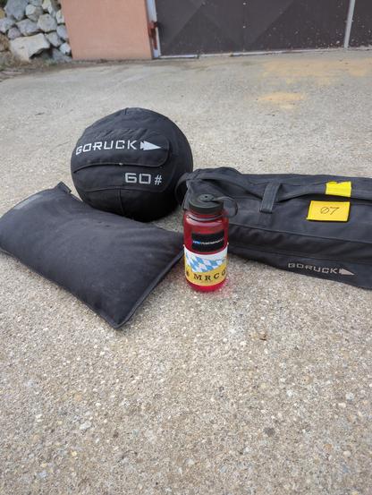 Three pieces of equipment from GORUCK: 60# medicine ball, 40# sandbag filler, 60# sandbag with 07 on its patch. They are behind a Nalgene bottle with a sticker that says MRC. All the equipment is on concrete.