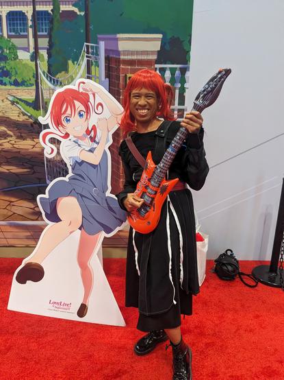Me in Mei Yoneme (CatChu outfit - black dress with guitar) cosplay next to Mei Yoneme cutout at Bandai Namco's booth.