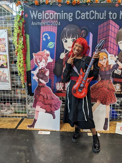 Me in Mei Yoneme (CatChu outfit - black dress with a guitar) cosplay in front of the Welcome CatChu banner and CatChu cutouts at ANYC.