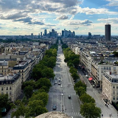 This photo shows a panoramic view of Paris, France. In the foreground, a wide tree-lined boulevard stretches toward the city center. The street is flanked by typical Parisian Haussmann-style buildings with their iconic cream-colored facades and mansard roofs. In the distance, the modern skyline of La Défense business district is visible, with its cluster of skyscrapers contrasting against the older architecture. The sky is dramatic, filled with a mix of puffy white clouds and patches of blue, creating a picturesque backdrop for the cityscape.