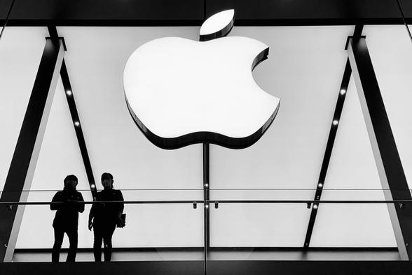 Auf einem S/W-Bild sind unter einem großen Apple-Logo die Silhouetten von zwei Personen auf einer Galerie zu sehen.