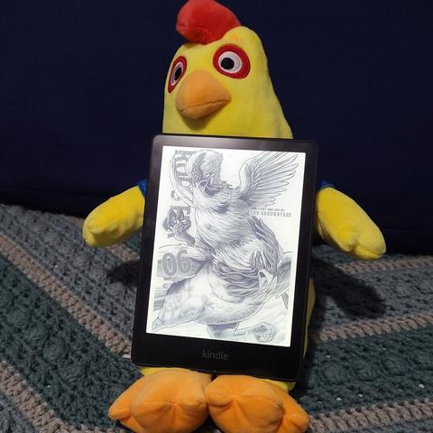 Photograph of a yellow, plush rooster toy laying on a blue quilt against a large blue pillow with a black framed eReader (Kindle) on its lap. The eReader has the screen opened to Rooster Fighter, Volume 6, which shows two, angry roosters charging toward the screen ready to fight.