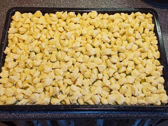 Ein Rhabarberkuchen in der Zubereitung, diesmal mit einer gleichmäßig verteilten Schicht von Streuseln bedeckt. Die Streusel sind aus hellgelbem Teig und bedecken den darunter liegenden Rhabarber vollständig. Der Kuchen befindet sich in einem rechteckigen Backblech und steht auf einer Küchenarbeitsfläche. Die grün-roten Rhabarberstücke, die zuvor sichtbar waren, sind jetzt fast komplett von der dicken Streuselschicht bedeckt.