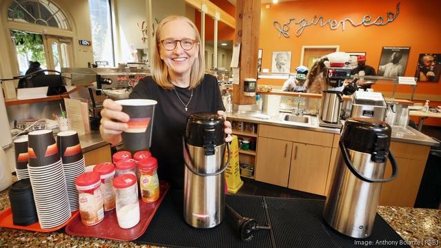 A smiling woman behind a coffee carafe offers the camera a cup of coffee.