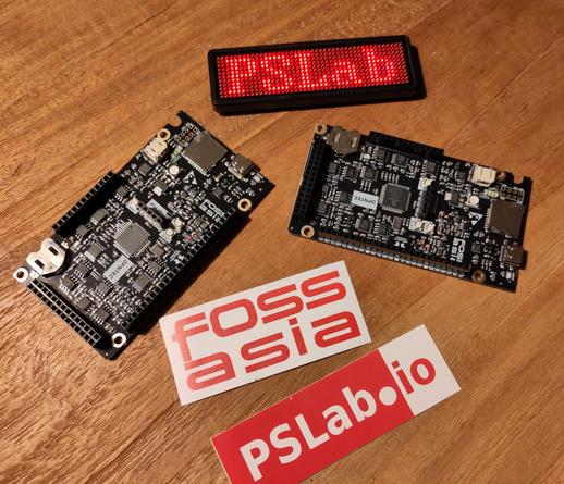 A bunch of stuff on a wooden table: Two PCBs, a little bit larger than an Arduino Uno with lots of headers. Also an LED badge which displays the text "P S Lab" and two stickers (one with the logo of Fossasia and one with the logo of PSLab.io).