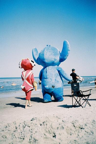 Photo of two cross-dressing plush characters on the beach. One of them is dressed as Stitch from Disney.