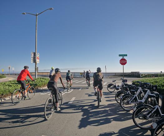 Lael Wilcox rides towards West Cliff Dr. in Santa Cruz CA. She is waving to fans who are waiting to see her ride through. She is followed by many riders, 5 of whom are visible, and many more are behind the camera.