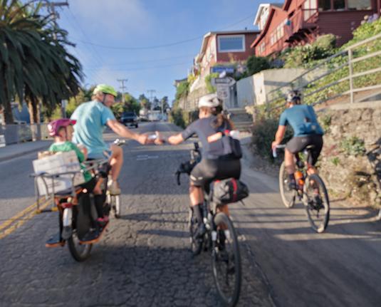 Lael Wilcox is handed a burrito wrapped in foil by a friend and fellow ex-Tour Divide racer. He is riding an orange cargo bike and has his son on the back of it. They are starting to climb out of Capitola CA. The photo is blurry because my camera had been accidentally nudged into manual focus mode and I didn't notice it yet.