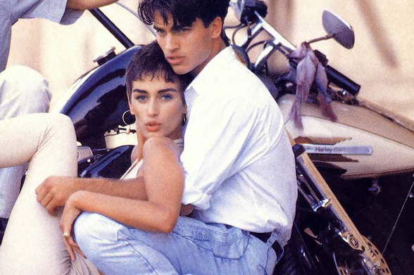 1990 Couple posing with a motorcycle.

"The image shows two individuals, likely a man and a woman, sitting together on a motorcycle.
The man is leaning forward slightly while holding onto the handlebars of the motorcycle, and both are looking directly at the camera.
The person in front appears to be wearing a white shirt with rolled-up sleeves and light blue jeans, while the one behind has short hair and is also dressed casually in similar attire.
They seem to be in a relaxed and candid pose, possibly posing for a photograph or just hanging out together."
Tags: "motorcycle, couple, two people, casual clothing, indoors, urban setting."