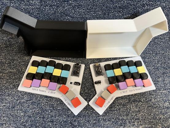 2 halves of a split keyboard arranged side by side. Has sculpted keycaps on the top and bottom row. In the background is a 3D printed travel case half printed in black PLA and half in white PLA