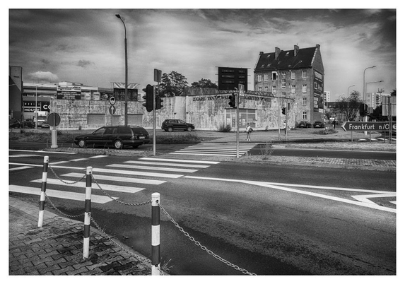 Strasseneinmündung mit Ampelanlage, Zebrastreifen, mehreren Fahrspuren, Sperrpfosten zwischen Fussgängerweg und Fahrbahn. Im Hintergrund einige Gebäude
