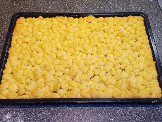 Auf einem schwarzen Backblech liegt ein rechteckiger Rhabarberkuchen mit einer goldgelben, krümeligen Streuseloberfläche. Darunter scheint der Kuchen leicht angebräunt zu sein. Die Streusel sind ungleichmäßig verteilt und variieren in ihrer Größe. Der Kuchen befindet sich auf einer dunklen Arbeitsfläche, die leicht bemehlt ist.