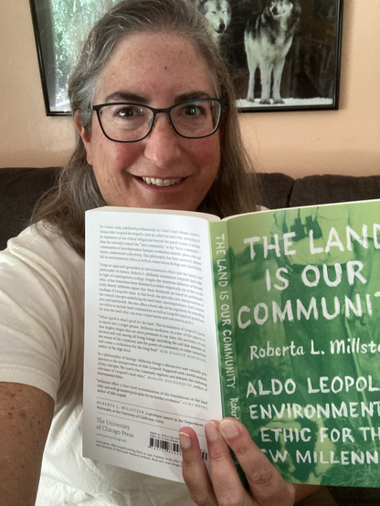 Silver haired author Roberta Millstein holding her book, The Land Is Our Community: Aldo Leopold's Environmental Ethic for the New Millenium.  There is a poster of wolves in the background.