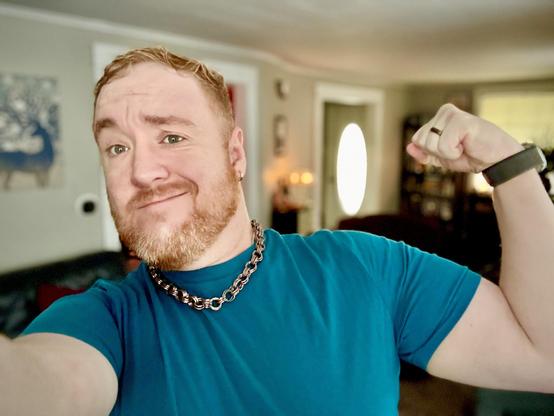 Mona Lisa-style oddly smirking bearded ginger man in a blue tshirt flexing for the camera.