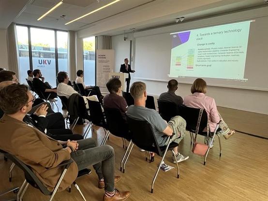 Das Bild zeigt einen Vortrag in einem modernen Konferenzraum. Ein Redner steht vorne am Pult und präsentiert eine Folie mit dem Titel "Towards a ternary technology stack" vor einer Gruppe von Zuhörern. Die Zuhörer sitzen in schwarzen Stühlen, die in Reihen aufgestellt sind, und konzentrieren sich auf den Vortrag. Im Hintergrund ist ein Roll-up mit der Aufschrift „Sichere Systeme“ zu sehen. Das Raumlicht ist hell, dank großer Fenster an der Seite, die Tageslicht hereinlassen. Die Atmosphäre wirkt professionell und fokussiert, passend zu einem Seminar oder einer Konferenz. Foto: Cyberagentur