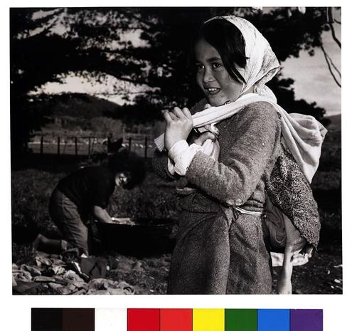 The image shows two individuals. One person is holding a child, possibly made of clothes or dressed in a way that makes it appear like a baby being carried. This person seems to be posing for the photograph, looking towards the camera with a smile. The other individual appears to be engaged in some sort of outdoor activity, likely washing clothes based on the context provided, and is turned away from the camera, focusing on their task.

The setting is a natural outdoor environment with trees and open space. There is no text present in the image to provide further information. The color palette consists of various shades of gray, white, black, and hints of green and yellow, suggesting that the photo may have been taken during daylight with natural lighting.

The image has a vintage feel, indicated by the clothing styles and the overall appearance of the photograph. It captures a moment of daily life, perhaps from an earlier time period, as suggested by the style of the attire.