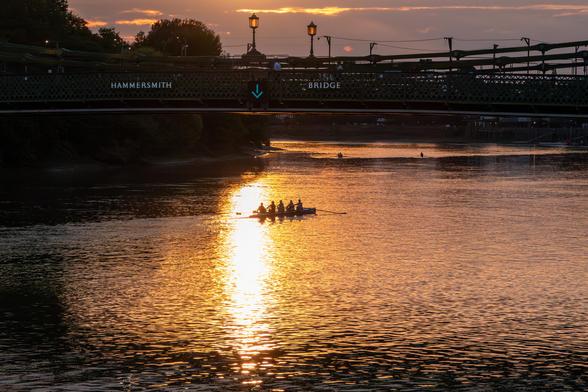 A sunset view under Hammersmith Bridge, there's an arrow pointing down on the bridge, directly beneath that is the sun reflected in a long line on the river, and people rowing a boat.