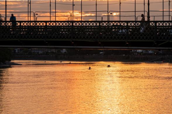 A sunset view under a bridge, one person walking on one side, the other leaning on the railing. The river below is golden with the sunset, and several rowboats are visible in the distance.