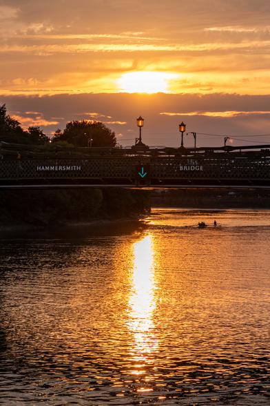 A sunset view under a bridge, one person walking on one side, the other leaning on the railing. The river below is golden with the sunset, and several rowboats are visible in the distance.