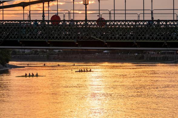 A sunset view under a bridge, with some people walking and others enjoying the view. The river below is golden with the sunset, and several rowboats are visible in the distance.