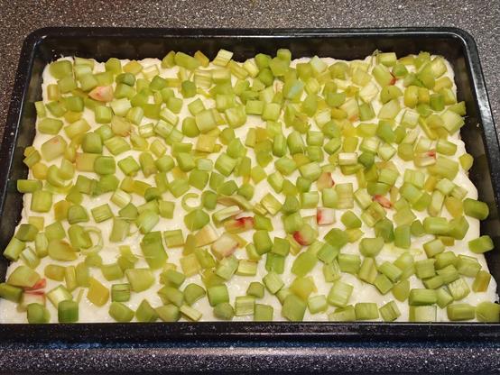 Ein ungebackener Rhabarberkuchen in einem rechteckigen Backblech. Der Kuchenboden ist mit einer dicken Schicht Pudding bedeckt, auf der gleichmäßig geschnittene grüne Rhabarberstücke verteilt sind. Die Rhabarberstücke sind teilweise auch rötlich gefärbt und liegen dicht beieinander. Der Kuchen befindet sich auf einer Küchenarbeitsfläche, bereit für den nächsten Zubereitungsschritt.