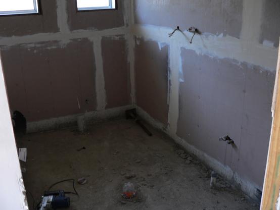 Looking through a doorway into a bathroom in the process of being renovated.  The floor is very rough concrete, plastic bottles blocking the drains, bare electrical wires hanging from the right wall, raw unpainted plasterboard on the walls. Loose concrete litters the floor, and a power tool can be seen at left.