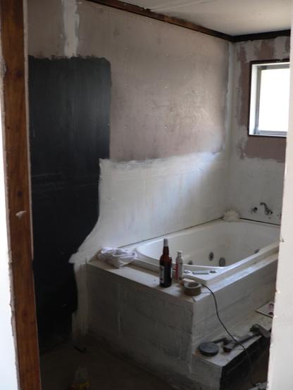 Looking through a doorway into a bathroom in the process of being renovated.  A spa bath is mounted in the rear left, with various building materials and tools lying about the room.  Waterproof membrane has been applied to the plasterboard around the bath and shower area at left.