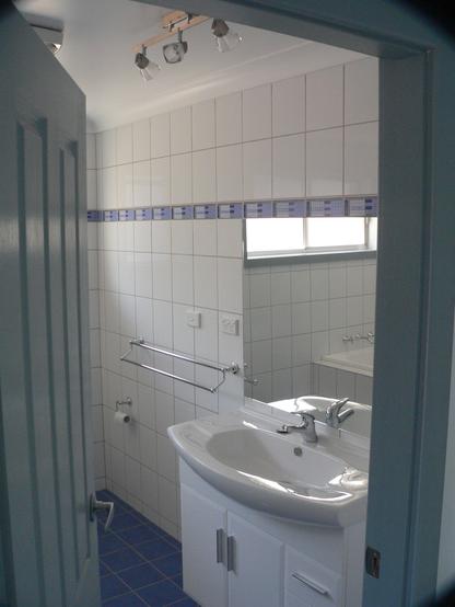 Same view as first image through the door looking right - this time of the renovated bathroom.  Sparkling white tiles cover the walls, a blue patterned frieze in one row at eye level, complementing the blue tiled floor. Glossy white vanity unit and a large frameless mirror at right, adjustable downlights above. Chrome towel rails further along the wall.