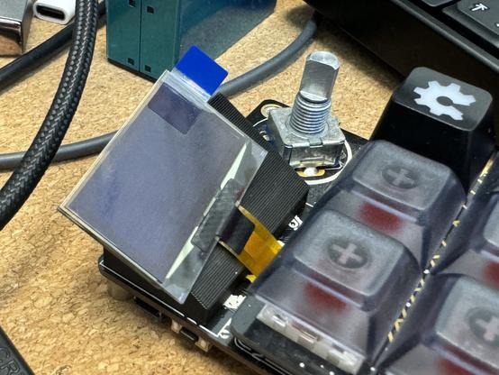 The front of the macropad, showing a new OLED screen with a protective film over the top and a blue tab for removing it. It has been positioned where the previous screen was, but on top of a small 3D printed wedge that tilts it forwards by 30 degrees towards the keys.