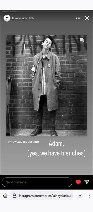 a black and white photo of a person with short dark hair in a trench coat standing in front of a brick wall with grafiti, sneering and lookong down at something.  overlay text reads Adam. (yes we have trenches) @newwavesocialclub