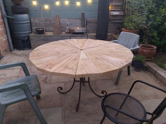 Photo of large round wooden table with spirally metal legs on a patio. There's three chairs around it and some pretty lights in background.