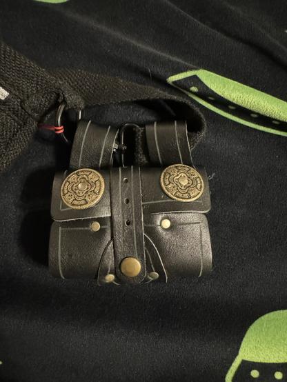 A small, black leather pouch with decorative circular clasps, resting on a dark fabric featuring green designs.