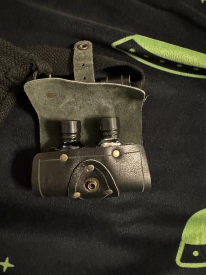 A dark green fabric background featuring a design with stars and a green object. On the foreground, there is a small leather case holding two black bottles, with metal details and a circular opening on the bottom.