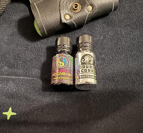 Two bottles of Double Scorpio poppers on a black blanket, double rainbow on the left and white gold on the right, above the bottles is a black leather pouch.
