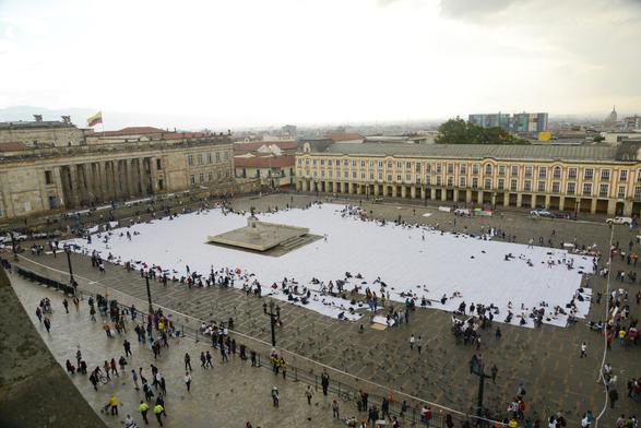 Вид на площадь Симона Боливара в Боготе с крыши общественного здания. Половину площади вокруг памятника занимают разложенные и частично сшитые одинаковые куски белой ткани. По краям копошатся десятки и десятки активист:ок. Пространство акции огорожено белой лентой, но часть публики зашла внутрь. // View of the Simon Bolivar Square in Bogota from the roof of a public building. Half of the area around the monument is occupied by identical pieces of white fabric laid out and partially sewn together. Dozens and dozens of activists are swarming around the edges. The space of the action is fenced off with white tape, but some of the public went inside. // Doris Salcedo, “Adding Absences” (“Sumando ausencias”), Plaza Bolivar, Bogota, 6–11 October 2016