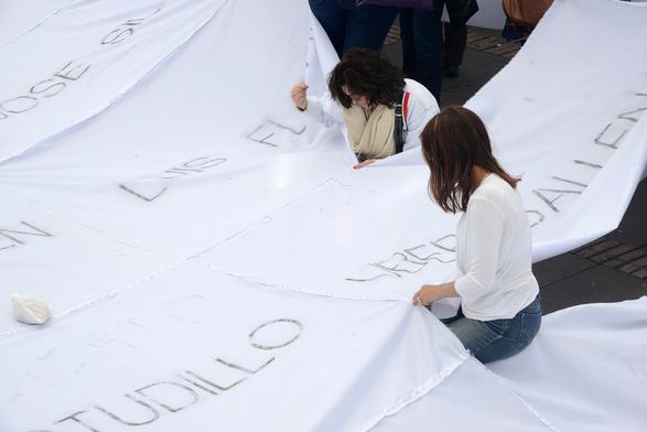 Две активистки с иголками в руках, снятые вблизи. Видно, что на сшитом полотне лежит мешочек-грузило, чтобы ветер не поднимал художественную работу высоко в воздух. В кадр поместились восемь отрезков ткани и пять имён пропавших без вести, все имена видны частично, ни одно имя целиком прочесть нельзя. // Two activists with needles in their hands, photographed close up. It is visible that a weighted bag is lying on the sewn canvas so that the wind does not lift the artwork high into the air. The frame contains eight pieces of fabric and five names of the missing, all names are partially visible, not a single name can be read in full. // Doris Salcedo, “Adding Absences” (“Sumando ausencias”), Plaza Bolivar, Bogota, 6–11 October 2016