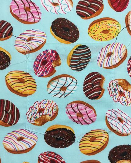 A pocket topstitched onto the front panel of a shirt. The tumbled pattern of frosted donuts on a cyan background, matches seamlessly