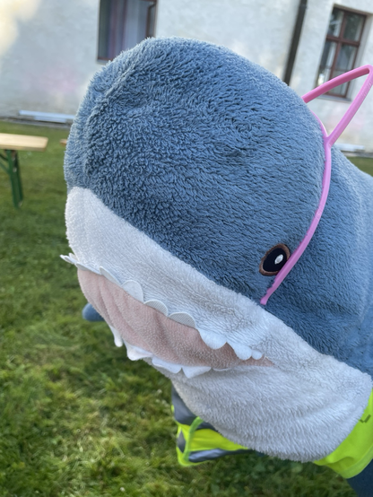 Blåhaj with Pink Cat Ears floating in the Air. In the Background is grass