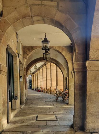 Soportales en la plaza de Cervantes de Santiago. Entre los soportales se pueden ver unas mesas y sillas. En primer plano hay unos arcos.