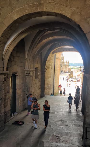 Pasaje entre la plaza del Obradoiro y la plaza de la Inmaculada. El pasaje está formado por arcos. Una gaitera toca música, mientras los turistas pasan a su lado.
