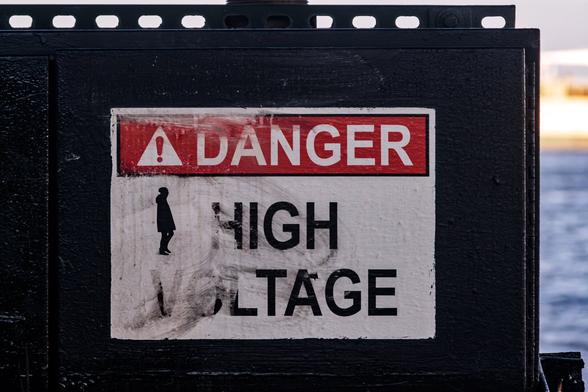 A color photograph of a warning sign. “Danger” in white letters on red along the top, and “High Voltage” below, black on white. On the left side of the sign, someone has added a black silhouetted walking figure facing towards the word “High”. Behind the cabinet the sign is mounted to, on the right side, the blue water of a river and a ship docked behind are visible, blurred, and lit by the late afternoon sun.
