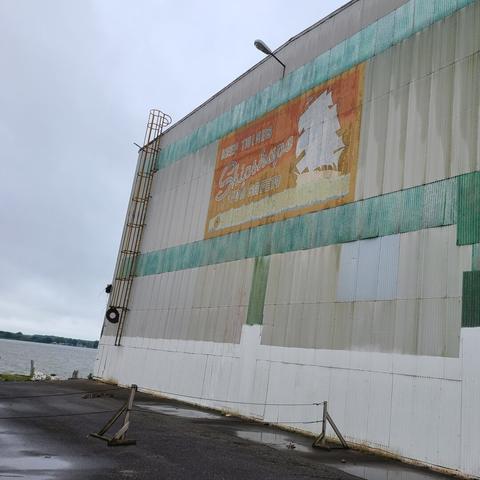 Photograph of a grey warehouse building with pale two green horizontal stripes, one near the middle and one near the top of the building. The building is adjacent to a small paved lot and waterfront can be seen in the background. Between the two pale green horizontal stripes is a faded sign with orange background and the outline of a large sailing vessel to the left. In large text, the remaining text on the sign reads, "Keep Things Shipshape for Safety." Beneath the text on the sign is a yellowish line which is unreadable.