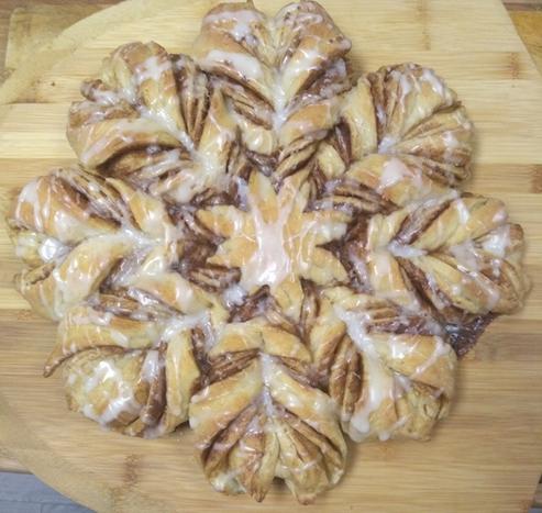 A cinnamon snowflake bread with 8  twisted, cinnamon swirled "arms" is drizzled with vanilla glaze. It measures 12" across and rests on a baking peel.