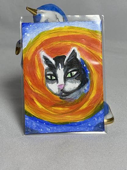 A vibrant watercolor painting of a cat's face, featuring green eyes and a black and white coat, set against a swirling background of orange and blue.