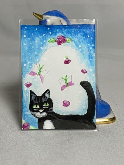 A painted card featuring a black cat with green eyes and a white patch on its chest, surrounded by flowers and a blue background. A decorative blue bird figurine is partially visible behind the card.