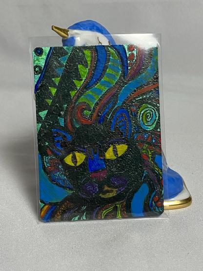 The image features an artistic card displaying a colorful depiction of a cat's face, characterized by bright colors and intricate patterns. In the background, there is a decorative object resembling a bird, with a blue and white design. The background is a light gray