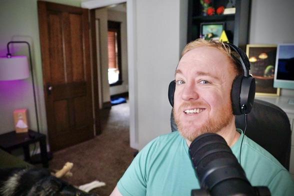 Bearded ginger man smiling at camera, headphones on and studio mic in front of him, giving the impression of a podcast or streamer
