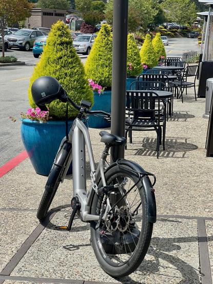 A silver-grey ebike is locked to a post. A black bicycle helmet is attached to its handlebars. In the background are some small evergreens and pink flowers; also, outdoor tables and chairs, all empty and available for use. At left and in distant background are many cars in parking lots.
