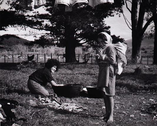 This is a black and white photo capturing a moment of domesticity in an outdoor setting. There are three people visible, each engaged in their own activities related to laundry day.

In the foreground, two young children are playing with clothes on the grass. They appear to be sorting or arranging the clothes, which are spread out before them. Their attire and the casual way they interact suggest a relaxed atmosphere, possibly during a family outing or camping trip where doing laundry has become part of their daily routine.

In the background, an adult woman is kneeling down with her back to us, engaged in a different task. She seems to be hanging clothes on a line strung between two trees. The act of hanging clothes suggests she might be preparing the laundry for drying or has just finished washing it and is now ready to let it air-dry.

The image captures a snapshot of life, highlighting the simplicity and beauty of everyday activities that are often overlooked in the fast pace of modern society. The lack of color adds a timeless quality to the scene, emphasizing the universality of these tasks across generations.