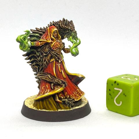 Miniature figure of a sorceress dressed in a red hood, bikini top, and skirt with flame designs around their edges. She holds a white orb in each hand that has green flames rising from them. A brown wyrm is wrapped around her back.